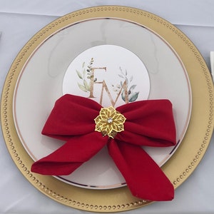 Op de afbeelding: Een gouden onderbord met een wit bord erop. Een rode strik met een gouden versiering is op het witte bord geplaatst. Een witte servet met gouden bestek ligt op de tafel naast het bord.