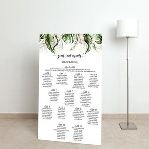 May include: A large white wedding seating chart with the text "your seat awaits" and names listed under table numbers. The top of the chart features a watercolor design of green leaves and pink accents. A modern floor lamp stands to the right.