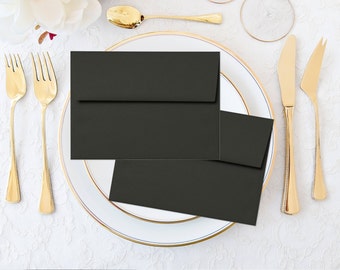 Può includere: Una vista dall'alto di una tavola formale con buste e biglietti neri su un piatto bianco con bordo dorato. Forchette, coltelli e cucchiai dorati sono disposti intorno al piatto su una tovaglia di pizzo bianco.