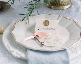 Può includere: Un piatto bianco con un bordo dorato, un nastro rosa legato attorno a una chiave vintage, una carta bianca con il testo "Boddie Whittaker" e un sigillo di cera, e un piccolo pezzo di carta con calligrafia.