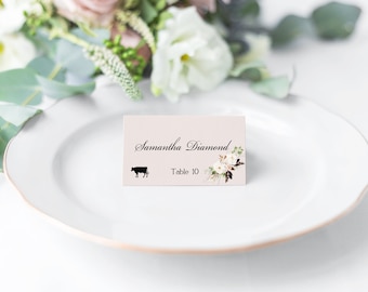 May include: A white place card with a pink background and floral design. The text on the card reads "Samantha Diamond, Table 10". A black silhouette of a cow is in the lower left corner.