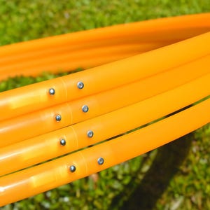 Puede incluir: Primer plano de aros de hula-hoop de color naranja brillante, parcialmente ensamblados, sobre un fondo de hierba verde. Los aros están hechos de plástico liso y están unidos con remaches plateados. La imagen destaca el color vibrante y el diseño sencillo.