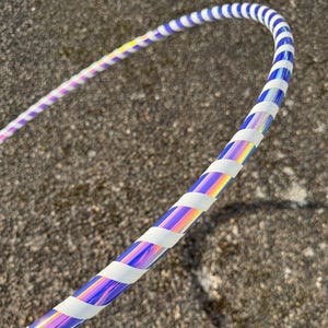 May include: A close-up of a colorful hula hoop. The hoop has a holographic, iridescent finish with blue and white stripes. White tape is wrapped around the hoop in intervals. The hoop is on a textured gray surface.