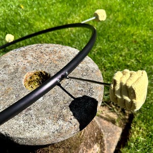 May include: A black metal fire hoop with two fire wicks, resting on a weathered stone surface. The wicks are made of a yellow, absorbent material. The background is a vibrant green lawn.