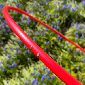 Puede incluir: Primer plano de un hula hoop rojo brillante. El aro está hecho de un material liso y sólido y está conectado con dos tornillos pequeños. El fondo es una vista borrosa de follaje verde y pequeñas flores moradas.