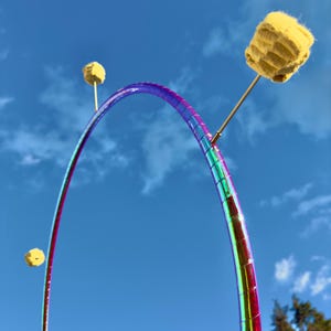 Puede incluir: Un aro iridiscente arcoíris con tres poi de fuego amarillos unidos. El aro está suspendido en el aire contra un cielo azul con nubes blancas.