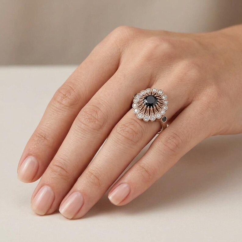 May include: A silver ring with a large, round, black gemstone at the center, surrounded by smaller white gemstones in a floral design. The ring is on a person's hand, showcasing the intricate details and elegant style of the jewelry.