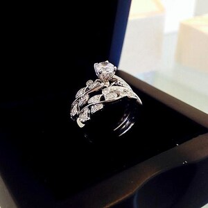 May include: A silver ring with a large, clear center stone and a delicate, leaf-shaped band. The ring is displayed in a black velvet box.