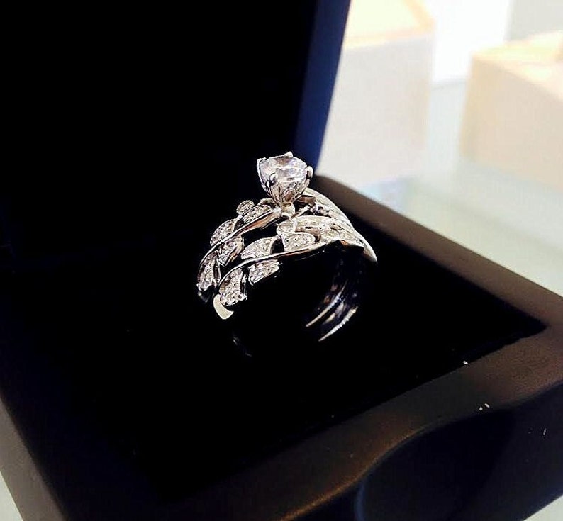 May include: A silver ring with a large, clear center stone and a delicate, leaf-shaped band. The ring is displayed in a black velvet box.