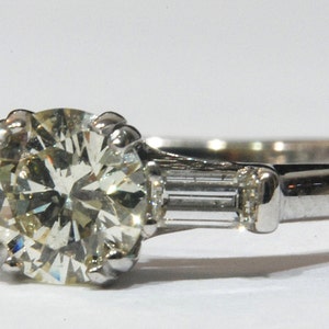 May include: A silver engagement ring with a large, round, clear diamond center stone and two smaller, rectangular, clear diamonds on either side.