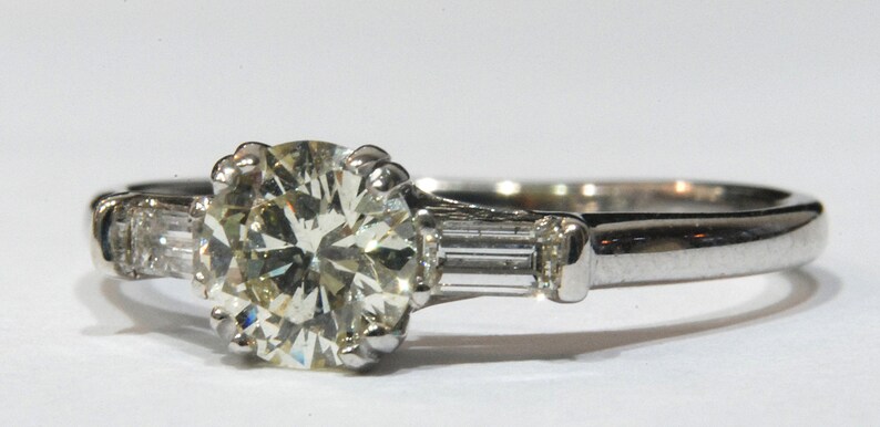 May include: A silver engagement ring with a large, round, clear diamond center stone and two smaller, rectangular, clear diamonds on either side.