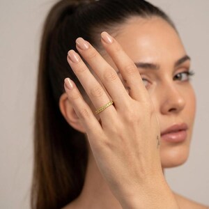 May include: A gold-colored ring with a continuous row of small, yellow gemstones. The ring is on a finger, with the hand and part of a person's face visible in the image. The ring appears to be a wedding band or eternity ring.