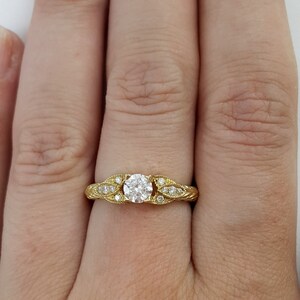 May include: A gold engagement ring with a round diamond center stone and smaller diamonds set in the band. The band is decorated with a leaf design.