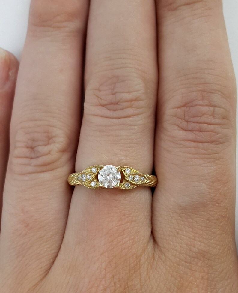 May include: A gold engagement ring with a round diamond center stone and smaller diamonds set in the band. The band is decorated with a leaf design.