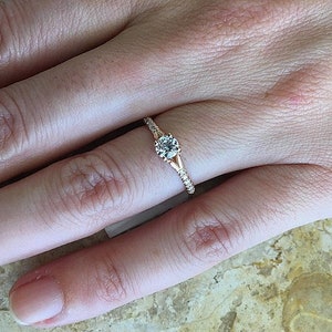 May include: A rose gold engagement ring with a single round diamond set in a prong setting. The band is adorned with small diamonds.