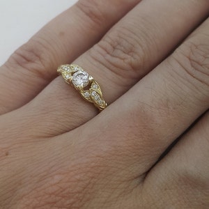 May include: A gold ring with a single round diamond set in a leaf-shaped design. The ring is set with smaller diamonds around the center stone.