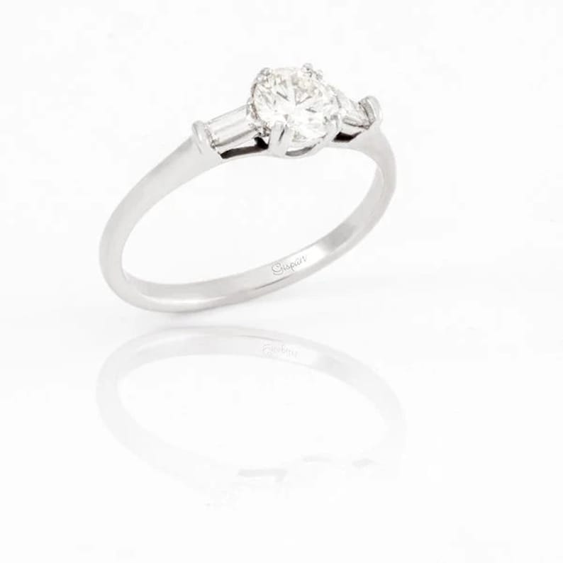 May include: A silver engagement ring with a round center diamond and two baguette diamonds on either side. The ring is engraved with the word "Gipar" on the inside band.