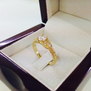 May include: A gold engagement ring with a single round diamond and smaller diamonds set in a delicate floral design. The ring is displayed in a velvet ring box.