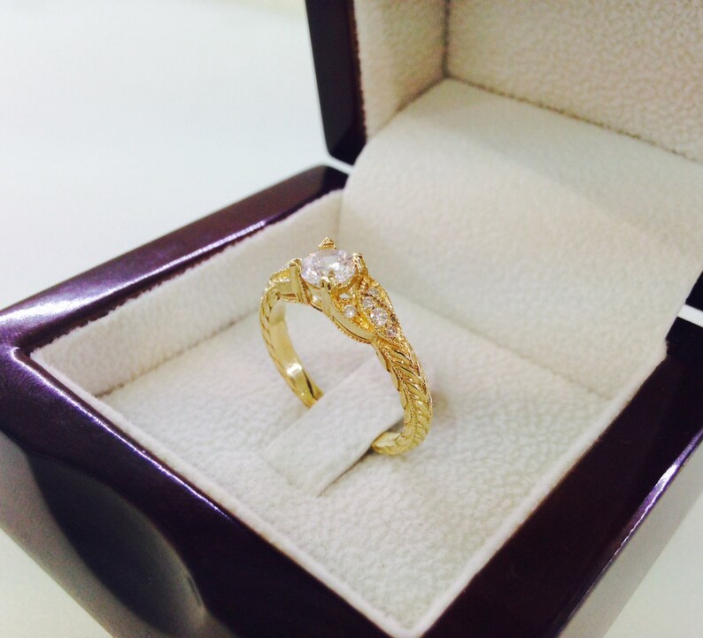 May include: A gold engagement ring with a single round diamond and smaller diamonds set in a delicate floral design. The ring is displayed in a velvet ring box.