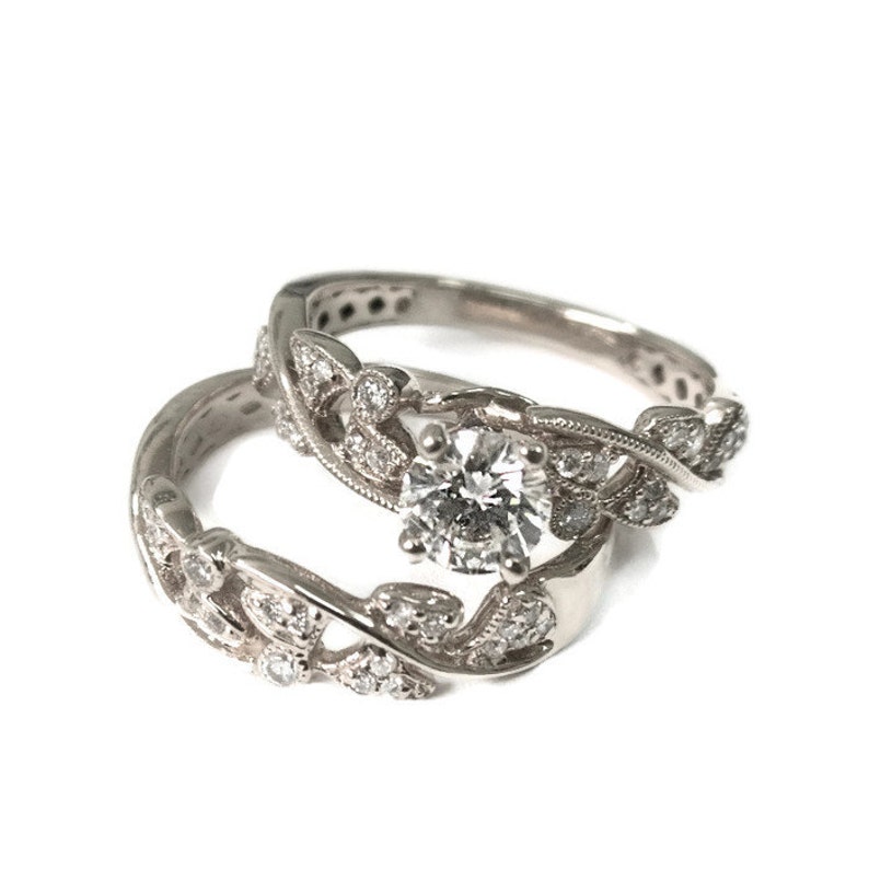 May include: A silver engagement ring set with a round diamond center stone and a delicate leaf design. The band is adorned with smaller diamonds.