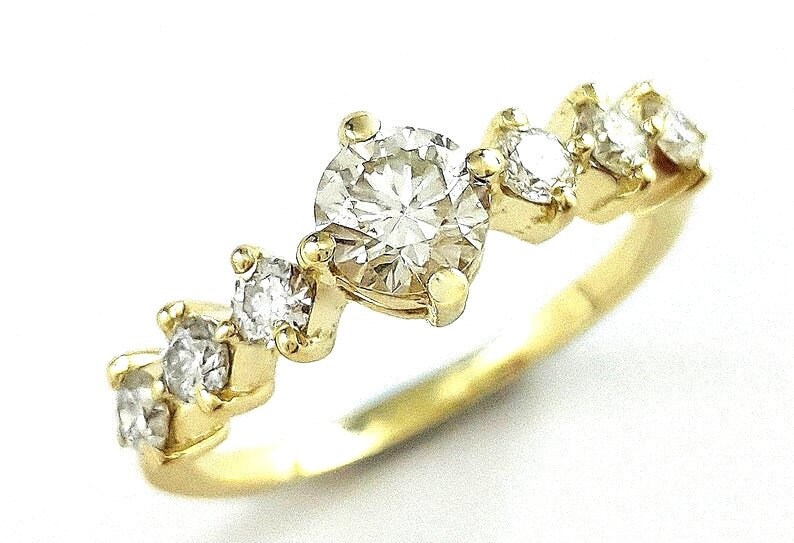 May include: A gold band ring with a row of five clear diamonds. The largest diamond is in the center and the other four diamonds are smaller and are on either side of the center diamond.