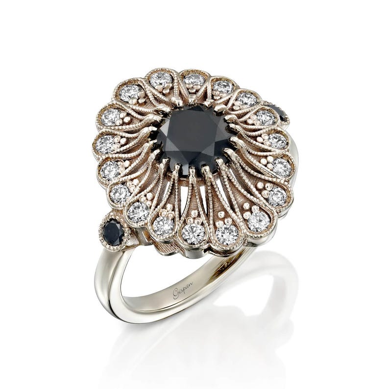 May include: A silver ring featuring a large, round black gemstone at the center, surrounded by smaller white gemstones. The ring has an ornate, floral-like design with a smaller black gemstone on the side. The ring is set against a white background.