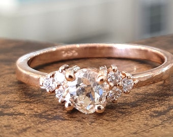 May include: A rose gold engagement ring with a large, round, clear center stone and smaller clear stones on either side. The band is textured.