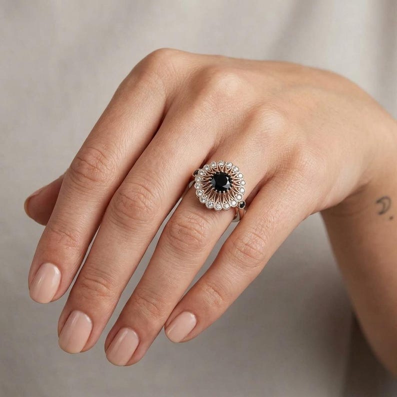 May include: A silver ring with a black center stone and a halo of small, clear stones. The ring has a floral design and is set on a hand with painted nails.