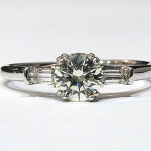 May include: A silver engagement ring with a large round center diamond and two smaller baguette diamonds on either side.