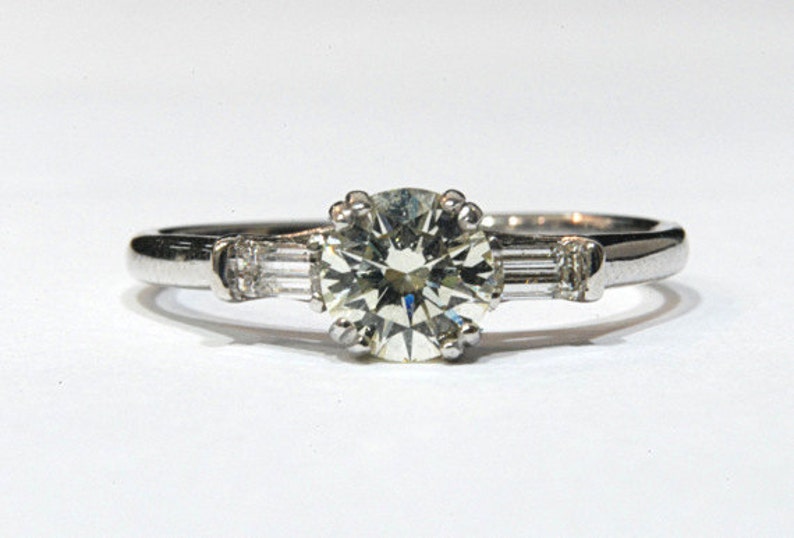 May include: A silver engagement ring with a large round center diamond and two smaller baguette diamonds on either side.