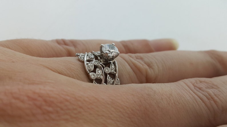 May include: A silver ring set with a large, round, clear diamond. The ring features a delicate, leafy design with smaller diamonds set around the main stone.