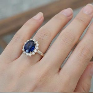 May include: A gold ring featuring a large, oval-cut, deep blue gemstone surrounded by a halo of smaller, clear gemstones. The ring is set on a delicate gold band, showcasing a classic and elegant design.