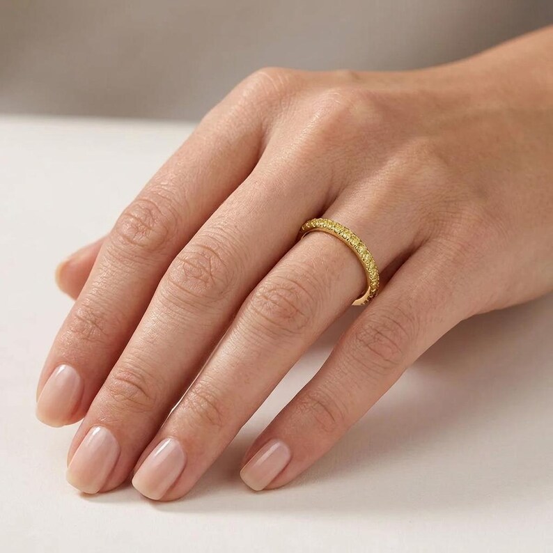 May include: A gold ring with a band of small, yellow gemstones. The ring is worn on a finger, showcasing its bright, polished appearance. The ring's design is simple yet elegant, with the gemstones set closely together.