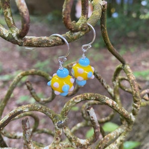 May include: Handmade earrings featuring yellow, blue, and pink glass beads. The earrings have silver hooks and hang from a weathered, rusty metal branch. The yellow beads are decorated with pink and blue spots.