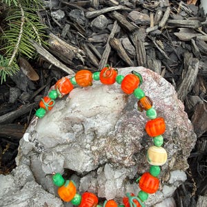 May include: A beaded bracelet featuring orange pumpkin-shaped beads, green beads, and a few off-white beads. The bracelet is strung on a silver chain and clasp. The beads are arranged in a repeating pattern, creating a festive accessory.