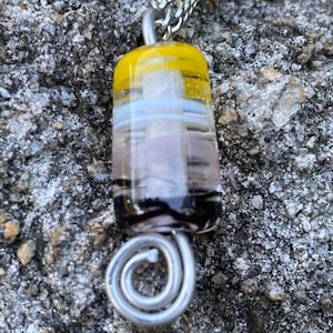 May include: A handmade glass pendant necklace with a silver chain. The pendant is a rectangular shape with layers of yellow, white, lavender, and black glass. The pendant is attached to a silver wire spiral.