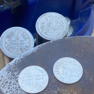 May include: Three silver coins with raised designs. The coins are on a dark gray surface. One coin has a cross with four circles around it. The other two coins have a profile of a person with a crown.