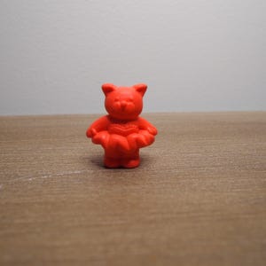 May include: A small, bright red bear-shaped figurine. The bear has a simple design with rounded features, including ears, arms, and a heart-shaped detail on its chest. It stands on a wooden surface against a plain white background.
