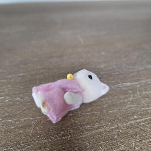 May include: A small, plush toy mouse wearing a pink dress. The mouse has a white body, a pink dress, and a yellow loop at the top. The toy is lying on a wooden surface.