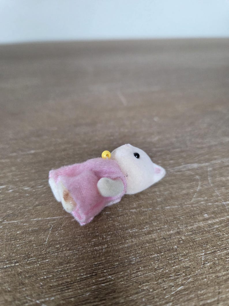 May include: A small, plush toy mouse wearing a pink dress. The mouse has a white body, a pink dress, and a yellow loop at the top. The toy is lying on a wooden surface.