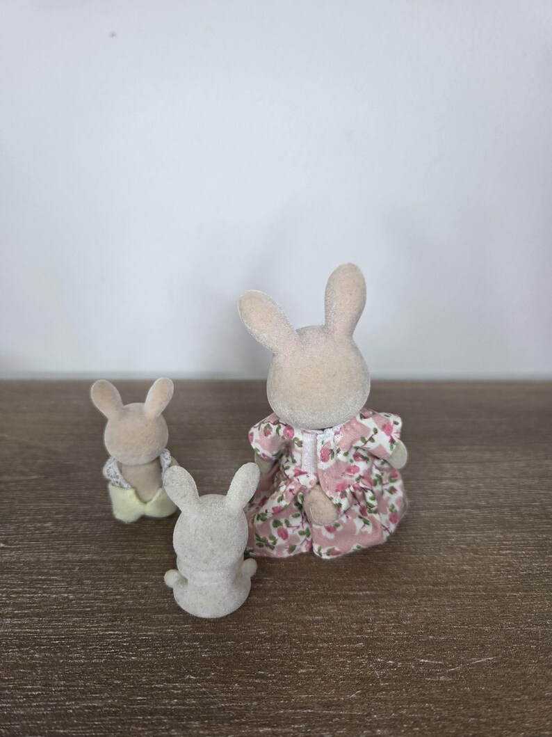 Pode incluir: Tr&ecirc;s pequenas figuras de coelhos. Um coelho est&aacute; vestindo um vestido floral rosa, outro est&aacute; vestindo uma camisa amarela e o terceiro &eacute; de cor cinza s&oacute;lida. Os coelhos est&atilde;o em uma superf&iacute;cie de madeira marrom.
