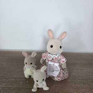 Pode incluir: Tr&ecirc;s figuras de coelhos de brinquedo. Um coelho adulto vestindo um vestido floral rosa e um avental branco. Dois coelhos menores est&atilde;o na frente. Os coelhos s&atilde;o bege claro com orelhas rosa e olhos pretos. O fundo &eacute; uma superf&iacute;cie de madeira clara e uma parede branca.