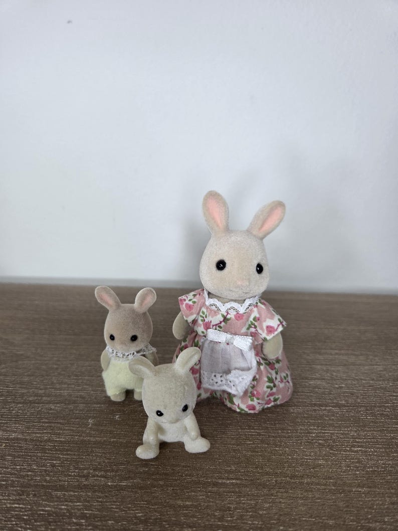Pode incluir: Tr&ecirc;s figuras de coelhos de brinquedo. Um coelho adulto vestindo um vestido floral rosa e um avental branco. Dois coelhos menores est&atilde;o na frente. Os coelhos s&atilde;o bege claro com orelhas rosa e olhos pretos. O fundo &eacute; uma superf&iacute;cie de madeira clara e uma parede branca.