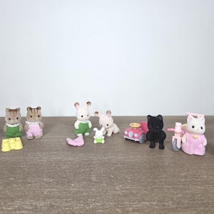 May include: A collection of miniature animal figurines, including squirrels, rabbits, and cats, dressed in colorful outfits. Accessories include a pink car and a tricycle. The toys are arranged on a wooden surface.