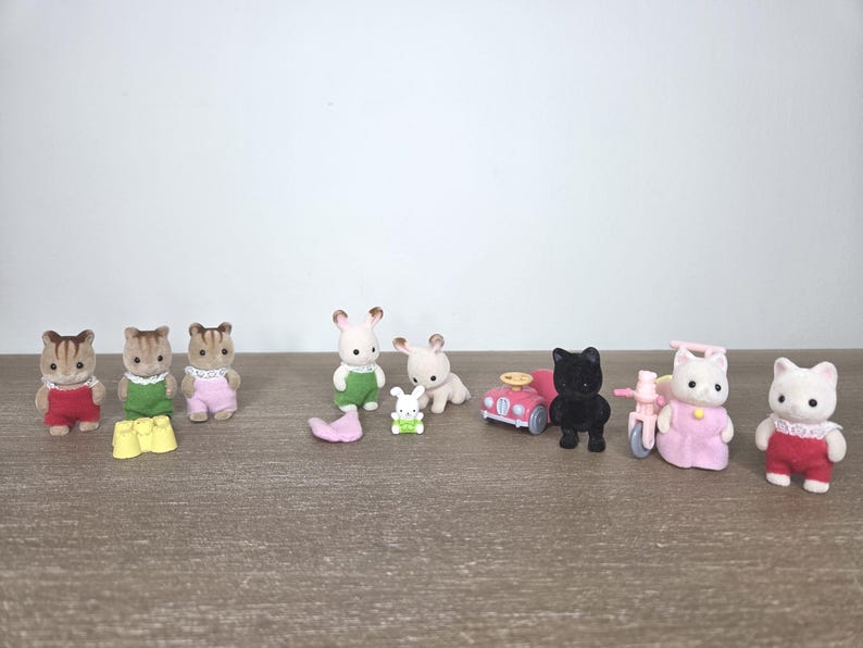 May include: A collection of miniature animal figurines, including squirrels, rabbits, and cats, dressed in colorful outfits. Accessories include a pink car and a tricycle. The toys are arranged on a wooden surface.
