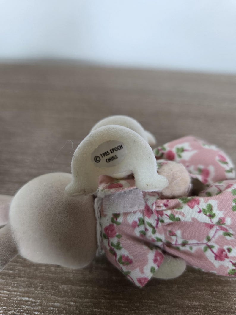 Pode incluir: Um coelho de brinquedo vintage com corpo rosa claro e orelhas brancas. O coelho usa um vestido floral rosa. O texto "1985 EPOCH CHINA" est&aacute; impresso na cabe&ccedil;a do coelho. O brinquedo &eacute; feito de material macio e felpudo.