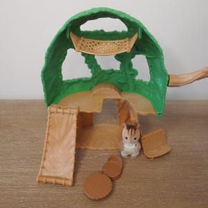 May include: A green and tan plastic playset resembling a treehouse, with a ladder, a small chair, and a toy animal. The treehouse has a green canopy with cut-out shapes and a tan base. The toy animal is a small, white and brown creature.