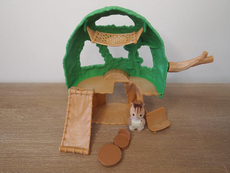 May include: A green and tan plastic playset resembling a treehouse, with a ladder, a small chair, and a toy animal. The treehouse has a green canopy with cut-out shapes and a tan base. The toy animal is a small, white and brown creature.
