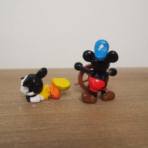May include: Two small figurines of cartoon characters. One is a black and white dog with yellow and orange accents. The other is a black mouse wearing a blue hat and red shorts, holding a brown steering wheel.