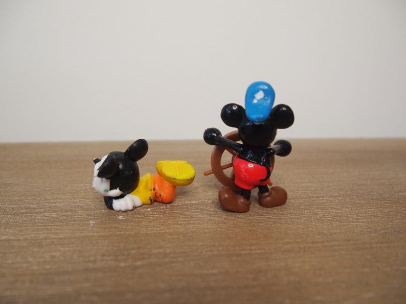 May include: Two small figurines of cartoon characters. One is a black and white dog with yellow and orange accents. The other is a black mouse wearing a blue hat and red shorts, holding a brown steering wheel.